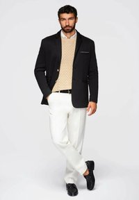 Schwarzer Blazer mit zwei Knöpfen über einem beigen Kabelstrickpullover, kombiniert mit weißen Hosen und schwarzen Schuhen, was ein elegantes Ensemble schafft.