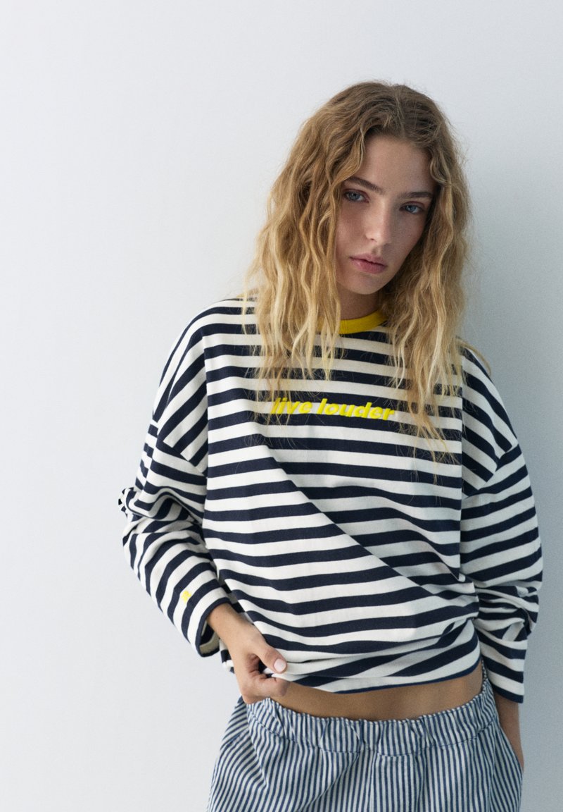 Jeune femme aux cheveux blonds ondulés portant un haut rayé noir et blanc avec l'inscription "live louder" et un pantalon rayé jaune et bleu.