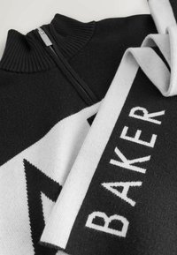 Sweater de punto en blanco y negro con cuello con cremallera, que presenta patrones geométricos audaces y la palabra "BAKER" destacada en blanco.