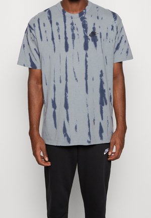 Una camiseta de color gris claro con un patrón de tie-dye en azul marino, mangas cortas, escote redondo y un pequeño logo negro de Nike en el pecho.