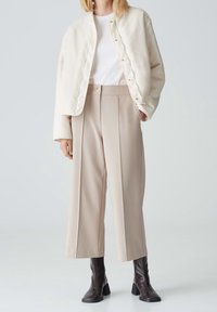 Giacca cropped beige chiaro, t-shirt bianca e pantaloni beige a gamba larga. Stivali marroni con tacco a blocco. Texture morbide e palette di colori neutri.