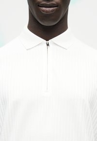 Gros plan d'une personne portant un polo blanc côtelé avec un col texturé et une fermeture éclair argentée partiellement zippée à l'avant.