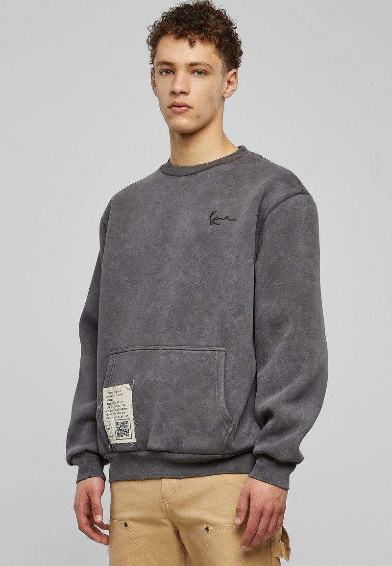 Felpa grigia con colletto tondo, maniche lunghe e tasca frontale. Presenta una finitura testurizzata e un'etichetta con dettagli stampati nella parte inferiore.