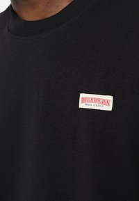 Čierne bavlnené tričko s okrúhlym výstrihom, ktoré má bielu a červenú nášivku "True Religion Brand Jeans" na ľavej hrudi.