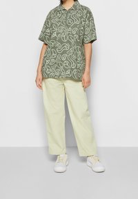 Polo verde a maniche corte con motivo di foglie bianche, abbinata a pantaloni a gamba larga giallo chiaro e scarpe da ginnastica bianche.