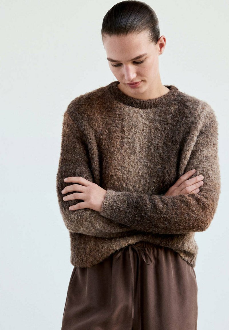 Personne aux cheveux foncés attachés porte un pull dégradé marron duveteux et un pantalon marron ample, debout, bras croisés et tête baissée.
