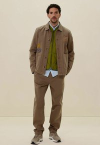 Bruine katoenen jas met geborduurde zon en golf, groene gewatteerde vest eronder, lichtblauw shirt, tan kleurige broek en grijze sneakers.