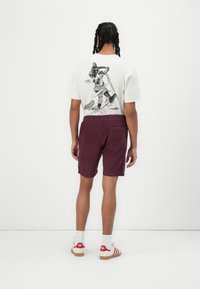 Witte t-shirt met een grafische print op de achterkant, gecombineerd met bordeauxrode shorts en roodgestreepte sneakers met witte veters, zichtbaar van achteren.