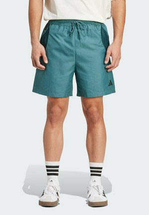 Teal elastiset shortsit, joissa on sivutaskut, kiristettävä vyötärö ja musta Adidas-logo. Yhdistetty valkoisiin kenkiin ja mustaraitaisiin sukkiin.