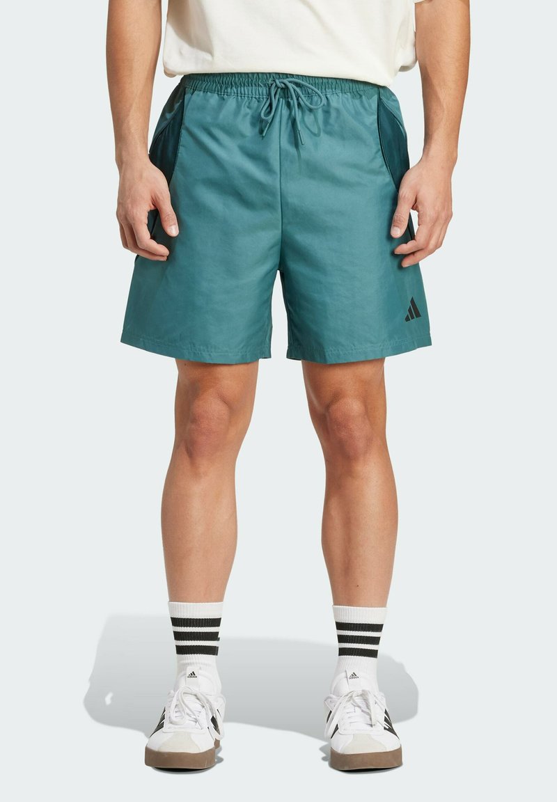 Turecké elastické šortky v morskej farbe so bočnými vreckami, sťahovacím pásom a čiernym logom Adidas. Nosí sa s bielymi topánkami a čiernymi pruhovanými ponožkami.