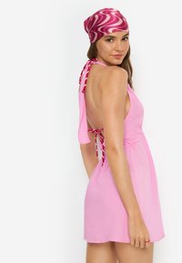 Ροζ φόρεμα σαντιγί με ανοιχτή πλάτη και λεπτομέρεια κορδονιού. Συνοδεύεται από ροζ και λευκό μαντήλι κεφαλής με σχέδιο. Μεταξένια υφή.