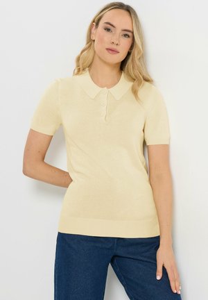 Femme aux longs cheveux blonds portant un pull polo à manches courtes jaune pâle et un jean bleu foncé sur fond blanc.