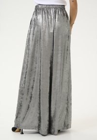 Jupe argentée, longue jusqu'au sol, fabriquée en tissu brillant et doux, avec une taille élastique. Texture lisse avec un reflets vertical et un design minimaliste.