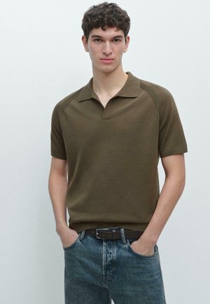Jeune homme aux cheveux bouclés portant un polo à manches courtes vert olive et un jean bleu, debout les mains dans les poches.