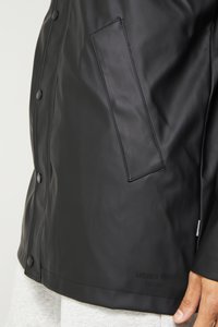 Black, smooth waterproof jacket featuring snap buttons, a patch pocket, and a logo printed near the hem. Soft gray fabric visible underneath.