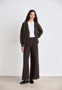 Bruine gebreide cardigan met een V-hals, knoopsluiting en losse mouwen, gecombineerd met wijde, donkerbruine broeken. Model draagt zwarte platte schoenen.