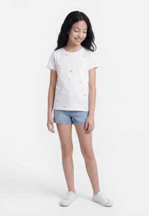 Pige med langt mørkt hår iført en hvid T-shirt med kirsebærprint, denimshorts og hvide sneakers, stående og kigger ned med et smil.