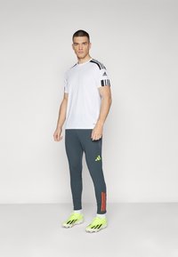 Bijela sportska majica s crnim prugama na ramenima, u kombinaciji s uskim tamnosivim hlačama s narančastim i neonsko zelenim naglascima. Svijetlo žuti kopački.