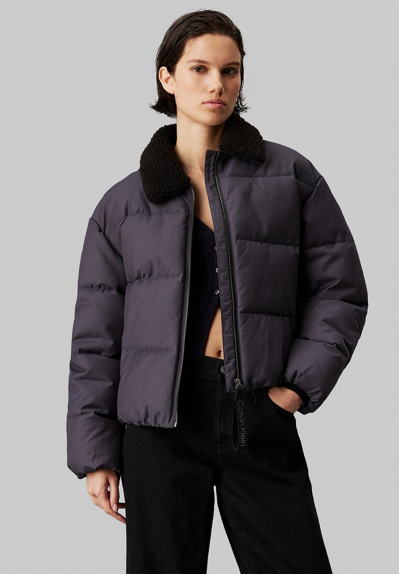 Pufferjack in donkergrijs met een zwarte fleece kraag, ritssluiting en een boxy pasvorm. Draag het over zwarte broek, met zichtbare gestikte secties.