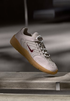 Svetlobéžová teniska s vínovočerveným logom Swoosh, bielymi šnúrkami a gumenou podrážkou, vznášajúca sa nad betónovým povrchom v jemnom osvetlení.