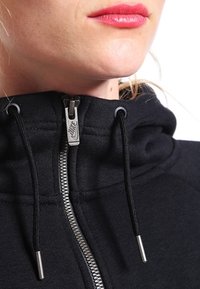 Svart zip-up hoodie med hög krage, silverfärgad dragkedja och svarta snören. Mjukt tyg med ingen synlig mönster eller logotyper.