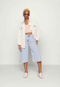 Mulher com cabelo curto loiro, vestindo um top beige, uma jaqueta oversized creme, culottes de denim claro, loafers bege com salto e óculos de sol, de pé.