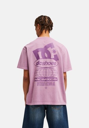 Osoba z zaplecionymi włosami ma na sobie fioletowy t-shirt DC Shoes z dużym logo, grafiką globusa i napisem MCMXCIV INTERNATIONAL na plecach.