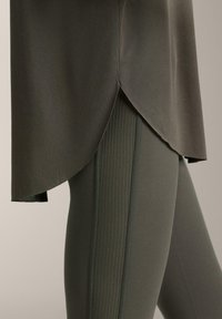 Gros plan sur un legging olive foncé avec un panneau latéral en tissu côtelé et un haut long assorti à ourlet courbé couvrant la cuisse.