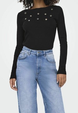 Mujer con camiseta negra de manga larga con ojales metálicos cerca del escote y vaqueros de mezclilla azul claro de talle alto.