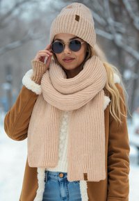 Beige strikket tørklæde og hat med ribbet tekstur. Hatten har et lille mærke; lagdelt med en tan-farvet frakke og en lys sweater. Solbriller bæres.