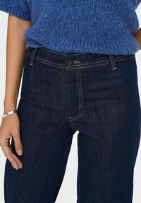 Tumši zili džinsi ar pieguļošu dizainu, kuriem ir šūta priekšējā kabata un metāla pogas aizdare. Virs tos redzams strukturēts zils adījums. 