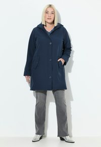 Impermeabile blu navy con cappuccio, bottoni frontali e due tasche laterali. Abbinato a jeans grigi e stivaletti d'argento. Tessuto liscio.