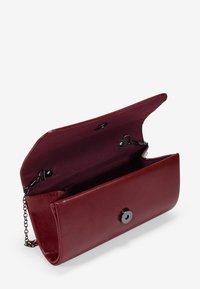 Clutch de imitación cuero burdeos con textura suave, cadena y cierre magnético. El interior presenta un forro oscuro y un clip.