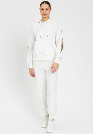 Giovane donna in piedi con felpa bianca con fessure aperte sulle maniche, pantaloni della tuta bianchi abbinati e scarpe da ginnastica bianche su sfondo neutro.