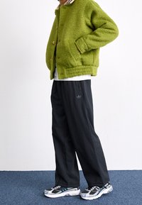 Grön fleecetröja med beige krage, svarta löst sittande byxor med en diskret logotyp och grå och vita löparskor.