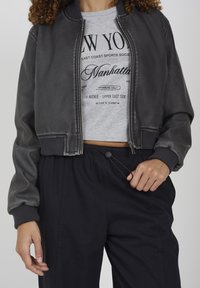 Persona che indossa una giacca bomber nera sopra un crop top grigio con la scritta "New York Manhattan", mentre aggiusta il cordoncino dei pantaloni neri.