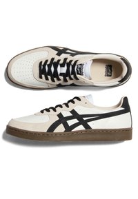 Białe i beżowe sneakersy wykonane ze skóry i zamszu, z czarnymi paskami akcentującymi, perforowanym noskiem i brązową gumową podeszwą.