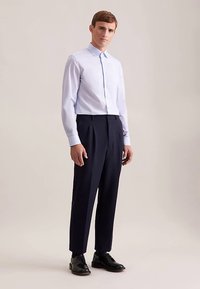 Lichtblauw overhemd met knopen en lange mouwen, marineblauwe geplooide broek, en zwarte schoenen. Gladde textuur en een getailleerde pasvorm versterken de silhouet.