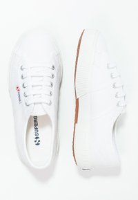 Sneaker bianche in tela con design con lacci, suole in gomma e dettagli testurizzati. Presentano una soletta con marchio e una suola esterna marrone a contrasto.