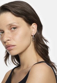 Silberne Creolen haben eine rechteckige Form mit einer strukturierten Oberfläche und hängen von einem kleinen gebogenen Steg. Das Model trägt ein schwarzes Tanktop.