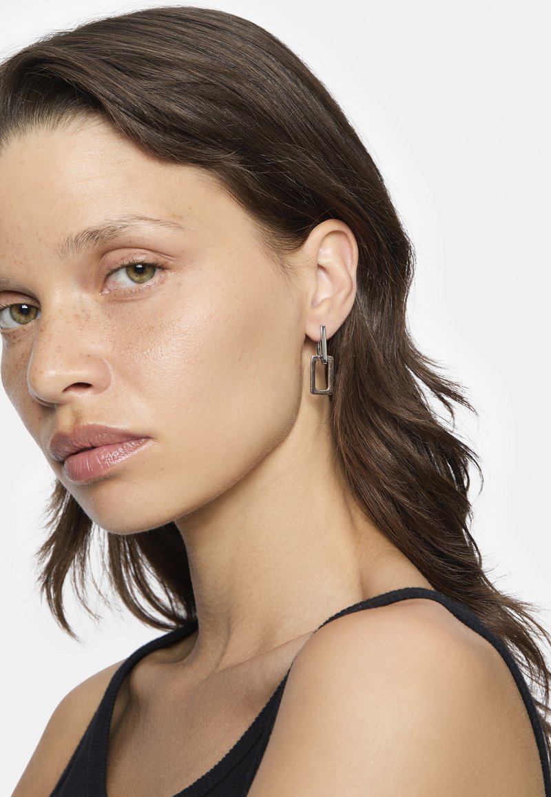 Silberne Creolen haben eine rechteckige Form mit einer strukturierten Oberfläche und hängen von einem kleinen gebogenen Steg. Das Model trägt ein schwarzes Tanktop.