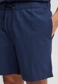 Granatowe szorty o teksturowanej powierzchni, z elastycznym pasem i sznurkiem do regulacji. Posiadają boczne kieszenie i prosty dół.