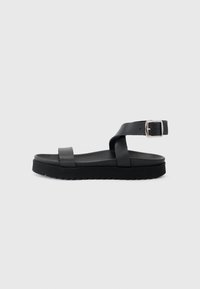 Sorte flate sandaler med en bred rem og en justerbar ankelrem med metallspenne, satt på en teksturert svart såle.
