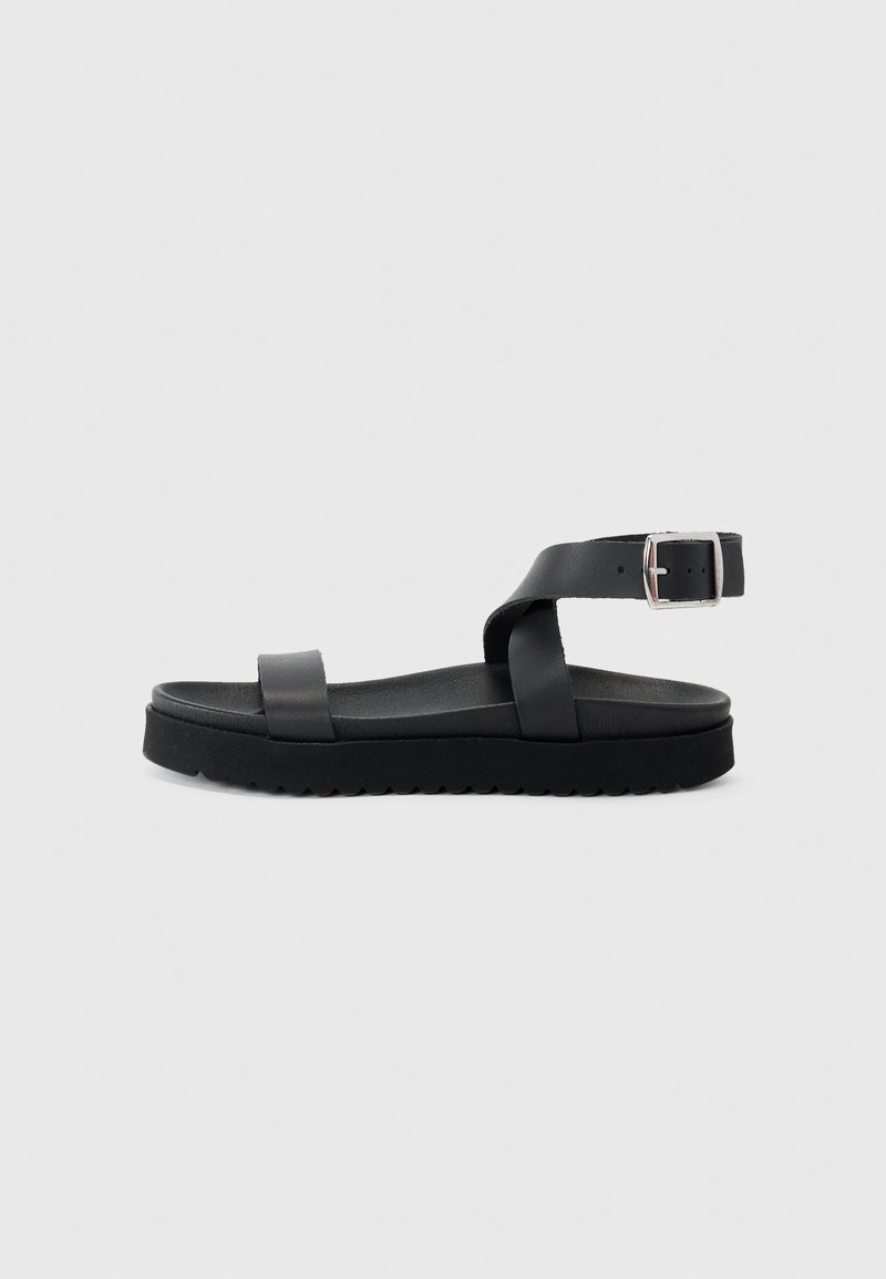 Sorte flate sandaler med en bred rem og en justerbar ankelrem med metallspenne, satt på en teksturert svart såle.