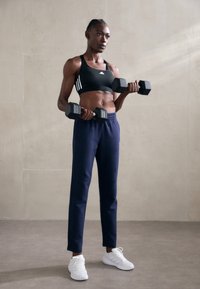 Athletic outfit featuring a black sports bra with three white stripes, navy tapered pants, and white sneakers, holding black dumbbells.