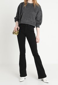 Sudadera gris corta, jeans negros acampanados, mules blancos de tacón y pequeño bolso amarillo a cuadros. Texturas suaves y fit casual.