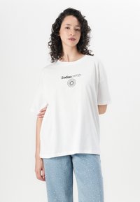 T-shirt de algodão branca com mangas curtas. Apresenta o texto impresso em preto "Energia Zodiac" e um design gráfico circular na frente.