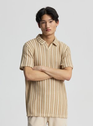 Giovane uomo con le braccia incrociate che indossa una polo in maglia a righe beige e bianche a maniche corte su uno sfondo chiaro e uniforme.