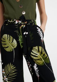 Svarta vida byxor med gröna tropiska blad- och blommönster; har en knytmidja och en subtil glans. I kombination med en grön knapp-up plagg.