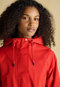 Kobieta z kręconymi ciemnymi włosami, ubrana w czerwony wodoodporny kaptur, zwrócona przodem z neutralnym wyrazem twarzy na beżowym tle.